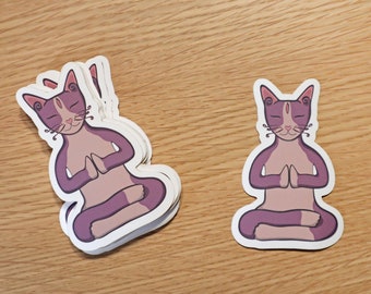 May include: A set of stickers featuring a purple cat meditating in a yoga pose. The cat has pink ears and a white patch on its chest. The stickers are on a wooden surface.