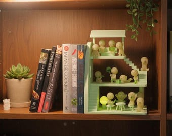 May include: A wooden shelf displays a succulent in a pot, several cookbooks, and a light green miniature house with small figurines. The cookbooks include titles like "Sushi Master" and "Japanese Cooking". A small white bear figurine is also present.