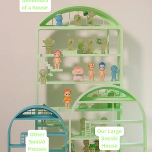 Puede incluir: Una exhibición de casas Smiski en varios tamaños y colores. La casa más grande es de color verde claro, con múltiples niveles y pequeñas figuras. Las casas más pequeñas son azules y verdes. El texto de la imagen dice "This behemoth of a house" y "Our Large Smiski House."