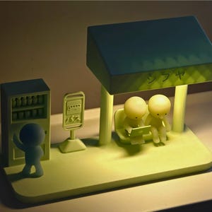 May include: A light green illuminated diorama lamp featuring a bus stop scene with a vending machine, a bus sign with Japanese text, and a shelter. Two figures sit on a bench under the shelter, while one figure stands by the vending machine.