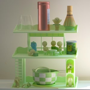 May include: A light green, three-tiered shelf unit displays tea ceremony items. It features a matcha whisk, a checkered bowl, a tin, and small figurines. The shelf is decorated with a dog and other figures.
