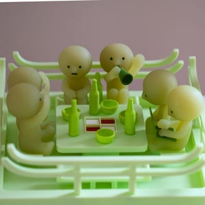 Può includere: Una scena in miniatura verde e gialla con piccole figure attorno a un tavolo. Il tavolo è apparecchiato con bottiglie e ciotole. Le figure hanno teste arrotondate e sono impegnate in varie attività.