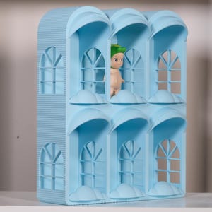 Op de afbeelding: Een lichtblauwe decoratieve structuur met meerdere ramen, gewelfde toppen en wolkvormige details. Een klein, naakt figuurtje met een groene hoed is zichtbaar in een van de ramen. De structuur heeft een gestructureerd, verticaal gestreept patroon.