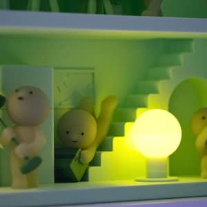 Puede incluir: Un diorama caprichoso con figuras en miniatura y elementos arquitect&oacute;nicos en tonos verdes y amarillos. Una figura sostiene una l&aacute;mpara, otra parece subir una escalera y una tercera toca una bocina. Un orbe brillante proporciona iluminaci&oacute;n.