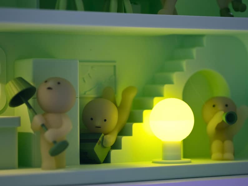 Puede incluir: Un diorama caprichoso con figuras en miniatura y elementos arquitect&oacute;nicos en tonos verdes y amarillos. Una figura sostiene una l&aacute;mpara, otra parece subir una escalera y una tercera toca una bocina. Un orbe brillante proporciona iluminaci&oacute;n.