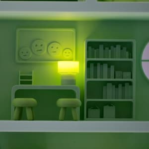 Puede incluir: Escena interior estilizada y monocrom&aacute;tica en tonos verdes. La habitaci&oacute;n presenta un escritorio con dos taburetes, una estanter&iacute;a con libros y una obra de arte mural con caras sonrientes. Una l&aacute;mpara brillante ilumina la escena, creando un ambiente suave y acogedor.