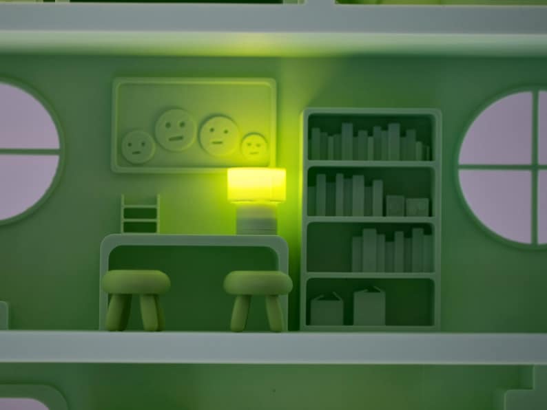 Puede incluir: Escena interior estilizada y monocrom&aacute;tica en tonos verdes. La habitaci&oacute;n presenta un escritorio con dos taburetes, una estanter&iacute;a con libros y una obra de arte mural con caras sonrientes. Una l&aacute;mpara brillante ilumina la escena, creando un ambiente suave y acogedor.