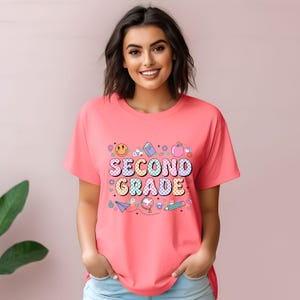 May include: A coral-colored t-shirt with the words "SECOND GRADE" in a colorful, checkered font. The design includes a smiling face, an apple, a book, a pencil, and other school-themed graphics. The shirt is a casual, short-sleeved style.