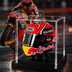Puede incluir: Un casco de motocicleta Shoei rojo, azul y amarillo con logotipos de Red Bull, Pull&Bear y Oakley, exhibido dentro de una vitrina de acrílico transparente sobre una base negra, con una imagen de fondo de un piloto de Repsol Honda.