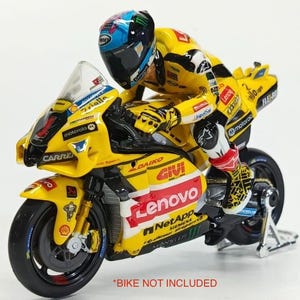 Może przedstawiać: Miniaturowa figurka motocyklisty w żółto-czarnym kombinezonie wyścigowym, w niebiesko-czarno-białym kasku, pochylona do przodu w pozycji wyścigowej. Figurka posiada różne logotypy sponsorów, w tym Lenovo, Givi oraz NetApp.