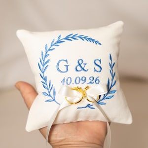 Peut inclure: Un coussin porte-alliances carré blanc avec des feuilles de laurier brodées en bleu, les lettres G & S, et la date 10.09.26. Deux anneaux en métal doré sont fixés au centre du coussin avec des rubans en satin blanc noués en nœuds.