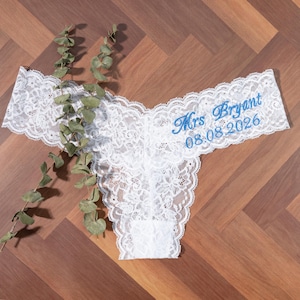 May include: White lace underwear featuring intricate floral patterns and scalloped edges. The fabric is embroidered with blue text that reads Mrs Bryant and the date 08.08.2026. A sprig of green eucalyptus leaves is placed across the left side.