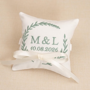 Peut inclure: Un coussin d'alliances carré blanc avec une broderie vert sauge représentant une couronne de laurier entourant les lettres M & L et la date 10.08.2026. Un ruban de satin blanc lisse est noué en boucle au centre du coussin brodé.