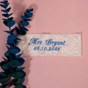 May include: A white lace garter with blue embroidered text that reads "Mrs Bryant" and "02.10.2026". The garter is set against a pink background, with blue-green leaves on the left side.