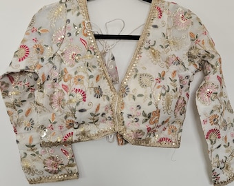 May include: A white long-sleeved blouse featuring a deep V-neckline and intricate floral embroidery in shades of pink, orange, and green. The garment is embellished with gold metallic thread and sequins, with a gold-trimmed border along the neckline.