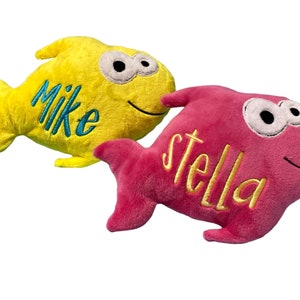 May include: Two plush fish toys with embroidered names. The yellow fish features the name Mike in blue thread, while the pink fish features the name stella in yellow thread. Both fish have large white eyes with black pupils and a stitched smile.