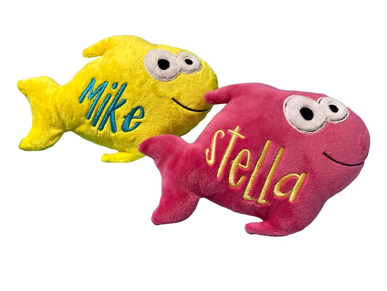 May include: Two plush fish toys with embroidered names. The yellow fish features the name Mike in blue thread, while the pink fish features the name stella in yellow thread. Both fish have large white eyes with black pupils and a stitched smile.