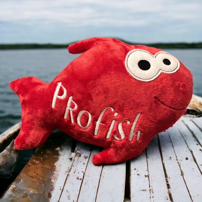 May include: A bright red plush fish toy featuring large white eyes with black pupils and the word "PROfish" embroidered in white thread across its side. The soft, stuffed fabric toy has a distinct fin shape and a simple stitched smile.