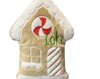 May include: A plush gingerbread house pillow in tan with white icing details on the roof and base. It features a red and white peppermint candy circular accent, a square window, and a striped door with the name Loki embroidered in green thread.