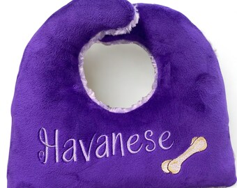 May include: A plush purple dog bib with a circular opening. The word "Havanese" is embroidered in cursive, along with a bone graphic. The bib has a soft, furry texture and a light-colored trim around the opening.