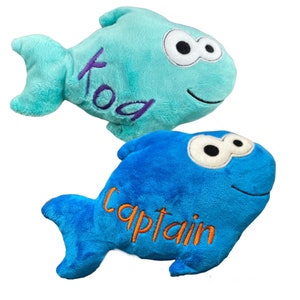 May include: Two plush fish toys with embroidered details. The top fish is light blue with the name "Koa" stitched in purple. The bottom fish is bright blue with the name "Captain" stitched in orange. Both feature large white eyes with black pupils.