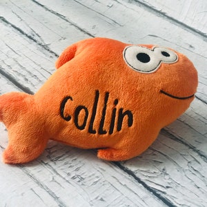 May include: A plush orange fish toy with a soft texture, featuring large white eyes with black pupils and a stitched black smile. The name Collin is embroidered in black thread on the side of the fish body, which has a fin and a tail.
