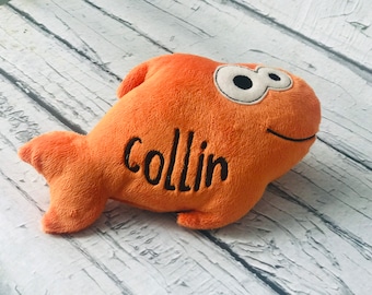 May include: A plush orange fish toy with a soft texture, featuring large white eyes with black pupils and a stitched black smile. The name Collin is embroidered in black thread on the side of the fish body, which has a fin and a tail.