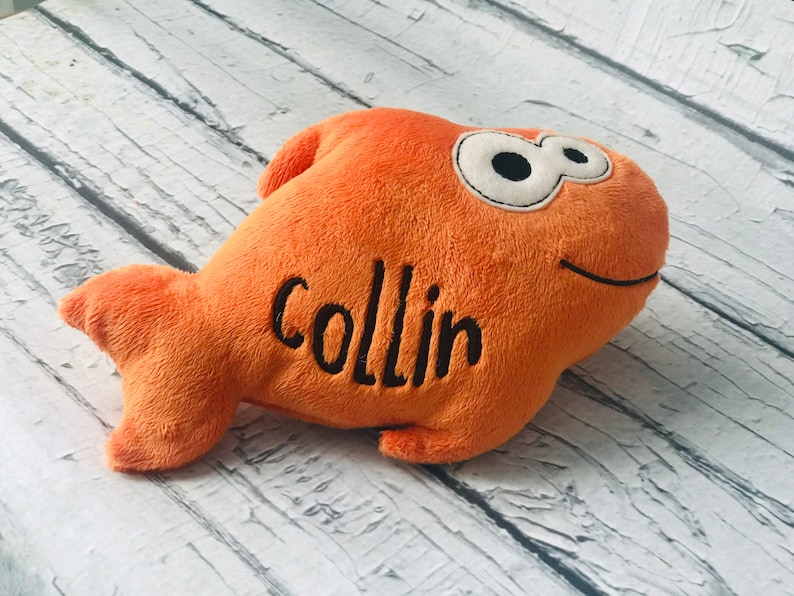 May include: A plush orange fish toy with a soft texture, featuring large white eyes with black pupils and a stitched black smile. The name Collin is embroidered in black thread on the side of the fish body, which has a fin and a tail.