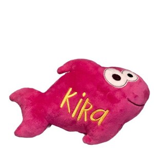 May include: A plush fish toy in a vibrant magenta color with a soft, fuzzy texture. The toy features large, white, oval-shaped eyes with black pupils and the name Kika embroidered in yellow cursive script on its side. It has a fin and tail.