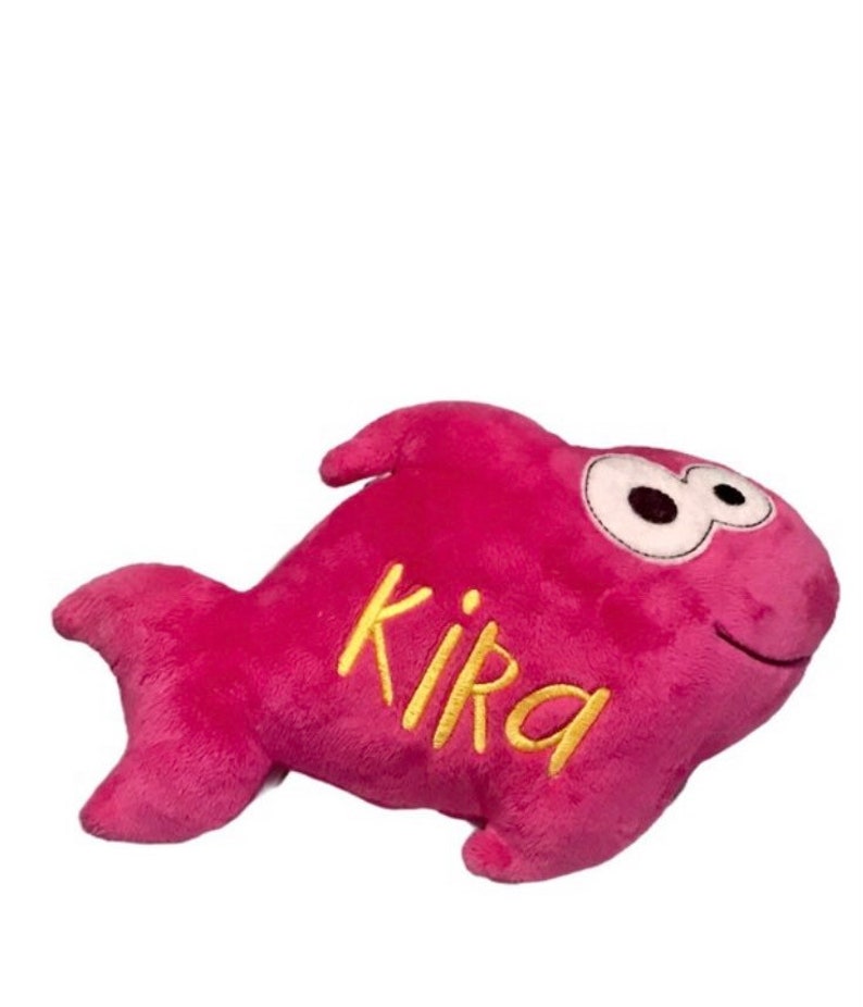 May include: A plush fish toy in a vibrant magenta color with a soft, fuzzy texture. The toy features large, white, oval-shaped eyes with black pupils and the name Kika embroidered in yellow cursive script on its side. It has a fin and tail.