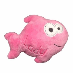 May include: A pink plush fish toy with a soft, fuzzy texture. The fish features large white eyes with black pupils, a stitched black mouth, and the word "lady" embroidered in pink thread on its side. It has a dorsal fin and a tail fin.