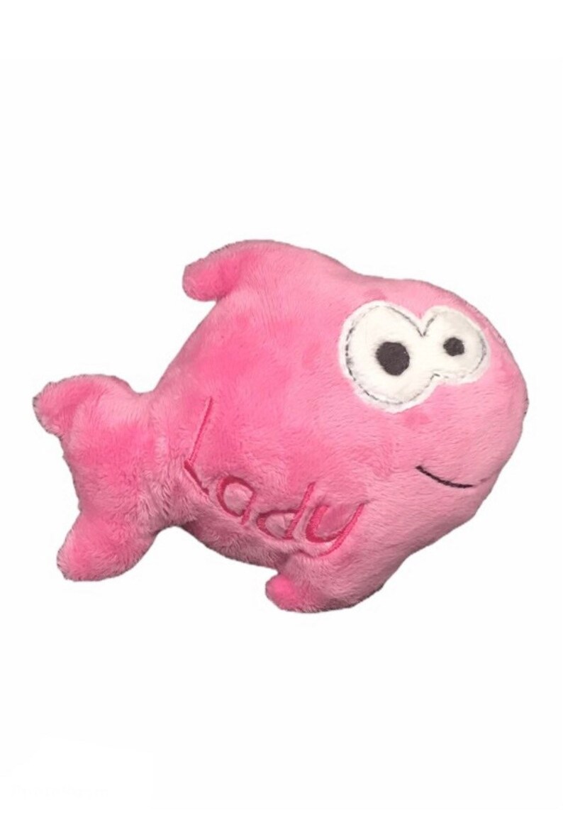 May include: A pink plush fish toy with a soft, fuzzy texture. The fish features large white eyes with black pupils, a stitched black mouth, and the word "lady" embroidered in pink thread on its side. It has a dorsal fin and a tail fin.