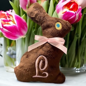 May include: A brown plush bunny rabbit with a pink bow and a pink letter L embroidered on its chest. The bunny is sitting in front of a vase of pink tulips.