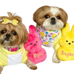 May include: Two Shih Tzu dogs dressed in yellow and white Easter outfits. One dog is holding a pink Peeps bunny with the name "Bijou" embroidered on it. The other dog is holding a yellow Peeps bunny with the name "Daphne" embroidered on it.