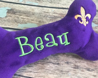 May include: A purple dog bone toy with the name "Beau" embroidered in green. A gold fleur-de-lis is embroidered on the top. The toy is made of plush material and is designed for pets.