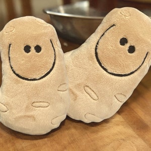 May include: Two plush brown potato toys with smiling faces. The potatoes are made of soft, fuzzy fabric and have a simple, embroidered design.