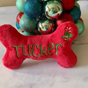 May include: Red plush dog bone toy with green embroidery that says "Tucker" and a candy cane with a green bow on the other side.