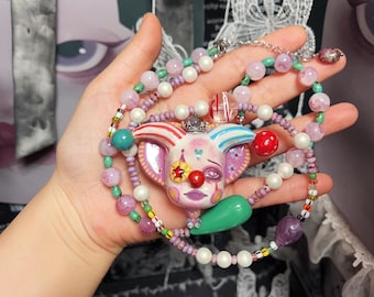 May include: A whimsical necklace featuring a clown pendant with a red nose, star, and colorful accents. The necklace includes pink, green, and white beads, with a silver chain. The clown pendant is the focal point of this unique jewelry piece.