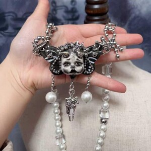 May include: A distinctive necklace with a central pendant depicting a painted face and horns, set against a black butterfly-wing design. The necklace incorporates a silver skeleton charm, pearl beads, and clear crystal elements, offering a gothic style.