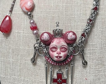 May include: A unique necklace featuring a pink and silver design. The pendant is a doll-like face with a crown, heart-shaped accents, and a red cross. The necklace includes a chainmail-style chain, beads, and dangling elements.