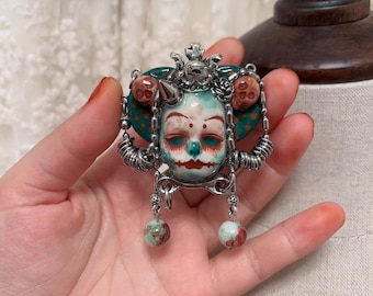 May include: A detailed, handcrafted brooch featuring a stylized face with a teal and white color scheme. The brooch has silver-toned metal accents, including coiled wire and dangling beads. The face has closed eyes and red accents.