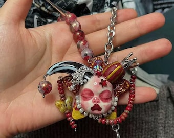 May include: A unique necklace featuring a central pendant with a doll face, crown, and jester elements. The pendant is adorned with red, white, and yellow accents, and is strung on a silver chain and beaded strand. A butterfly and star add to the whimsical design.