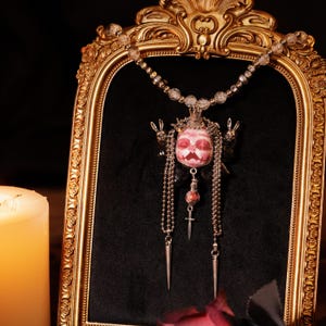 以下が含まれることがあります： ピンクと白の頭蓋骨のデザイン、王冠、ぶら下がっているチェーン、小さな十字架が特徴のユニークなペンダントネックレス。 ネックレスは、黒い背景に対して、金色の装飾フレーム内に表示されます。