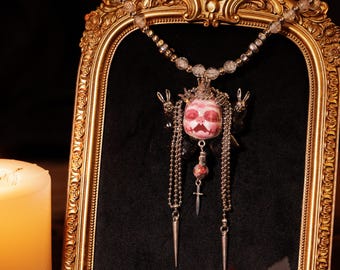 May include: A unique pendant necklace featuring a pink and white skull design with a crown, dangling chains, and a small cross. The necklace is displayed within a gold ornate frame against a black backdrop.