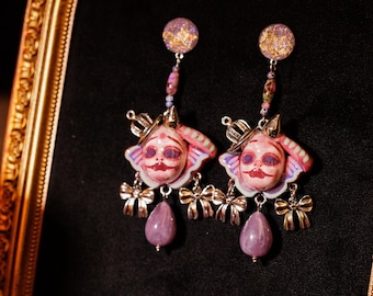 May include: A pair of dangle earrings featuring a circular stud with gold flakes, a small bead, and a central charm shaped like a face with a crown and wings. Two silver-toned bows hang from the sides, and a smooth lavender teardrop bead hangs below.