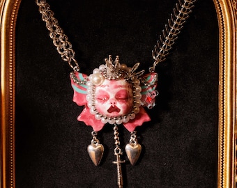 May include: A silver-toned necklace featuring a pendant with a doll-like face, a crown, and a pink bow. The pendant is adorned with pearls and red accents. Two heart-shaped charms and a syringe-shaped charm dangle below.