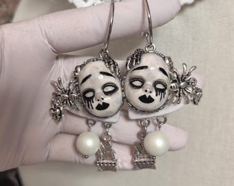 May include: A pair of silver hoop earrings featuring white doll face charms with black painted eyes and lips, accented by silver spider charms, small white pearl-like beads, and miniature silver castle charms hanging from the bottom of each face.