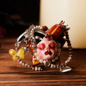 May include: A whimsical brooch featuring a central, stylized face with closed eyes and a red mouth, surrounded by pearls and a beaded border. The brooch includes a butterfly, a crown, and a black and white striped element.