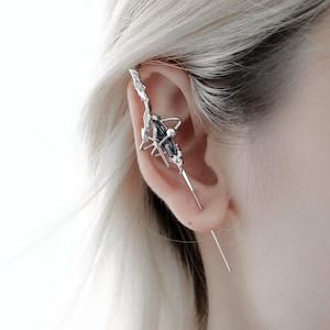 Puede incluir: Un ear cuff plateado con una piedra preciosa gris oscuro y un diseño único y puntiagudo. El cuff se envuelve alrededor de la oreja, con un elemento largo y puntiagudo que cuelga debajo del lóbulo. La joya tiene una estética vanguardista y moderna.