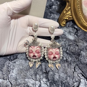 Könnte beinhalten: Ein Paar verzierter Ohrringe mit einem Puppengesicht-Design. Jeder Ohrring hat ein perlenbesetztes Gesicht mit geschlossenen Augen, einer Krone und baumelnden silbernen Tropfenanhängern. Die Ohrringe sind mit Perlen und silbernen Details verziert.